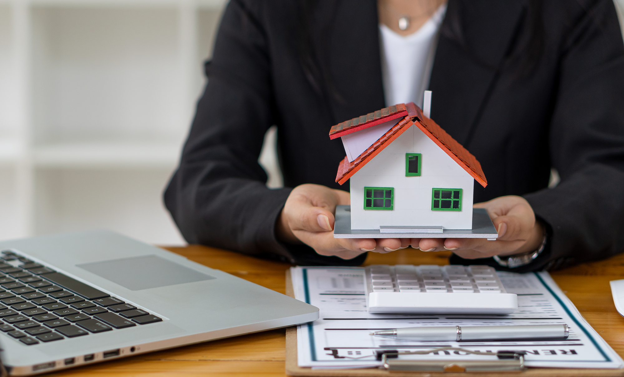 A woman holding a model of a house in front of a laptop.