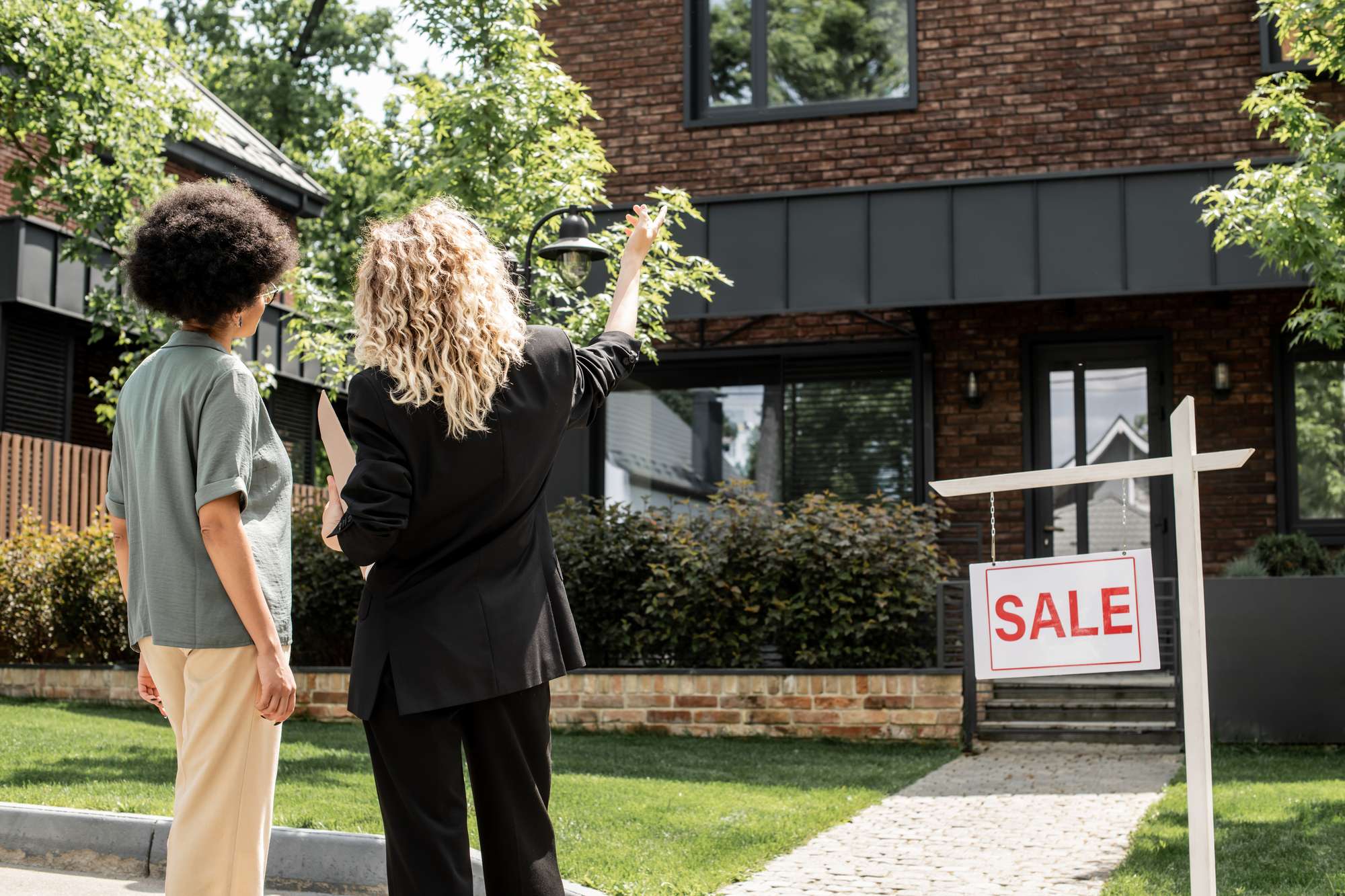 Two women standing in front of a house with a sale sign.