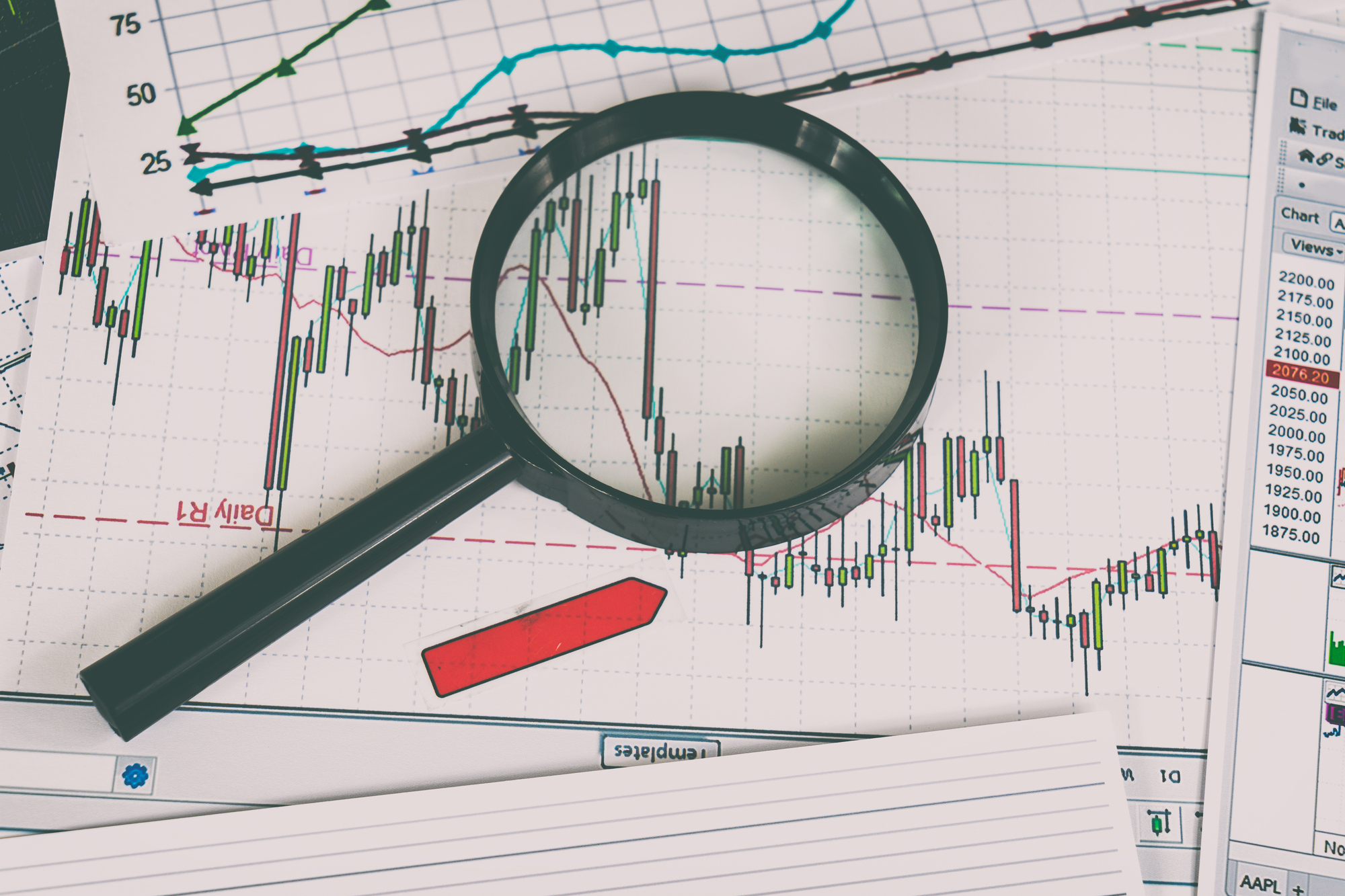 A magnifying glass is on top of a sheet of paper with stock charts.