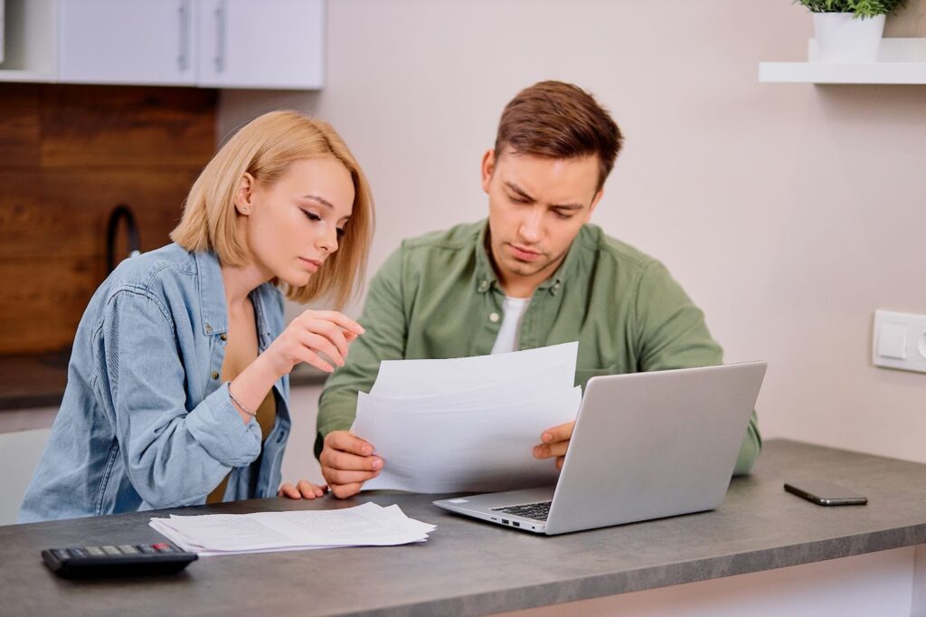 A man and woman are looking at paperwork on a laptop.
