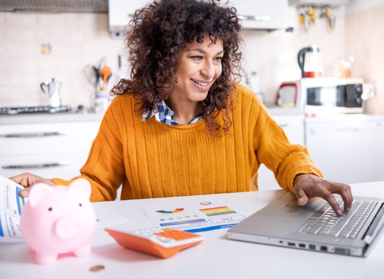 A woman is working on her laptop while holding a piggy bank, possibly saving up for real estate investments.