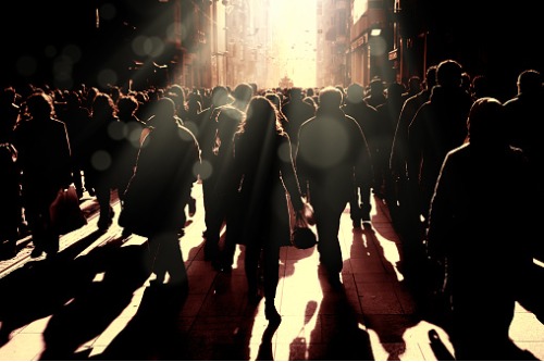 A crowd of people walking down a street with the sun shining on them.