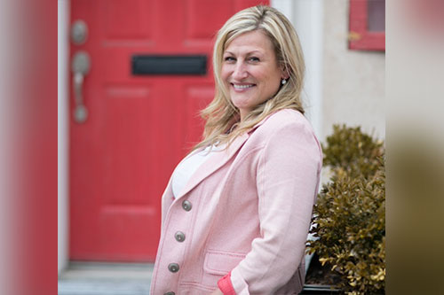 A woman in a pink blazer standing in front of a red door.
