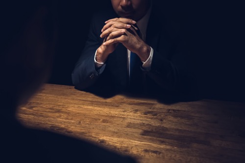 A man in a suit sits at a table with his hands clasped.