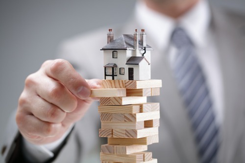 A businessman holding a model house on top of a stack of wooden blocks.