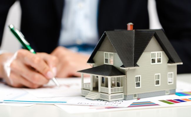 A woman in a business suit is holding a model of a house and a pen.
