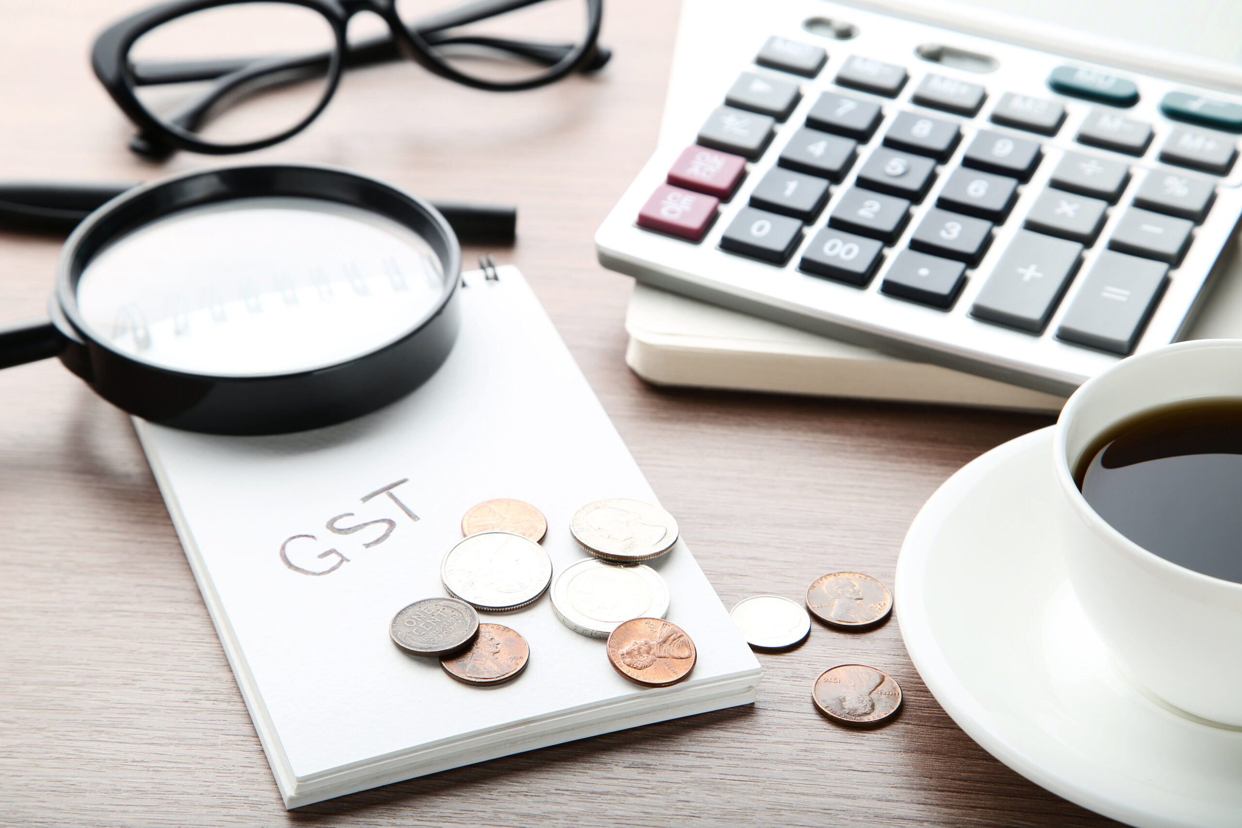 Inscription GST on paper with coins, magnifying glass, calculator and cup of coffee