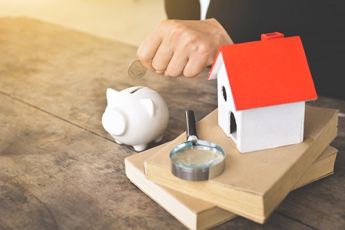 A hand holding a magnifying glass and a piggy bank next to a house.