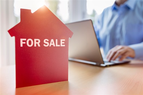 A man working on a laptop with a house for sale sign.