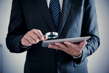 A businessman holding a magnifying glass while using a tablet.