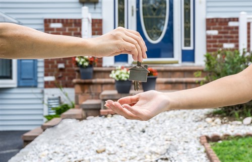 A woman handing a house key to another person in front of a house.