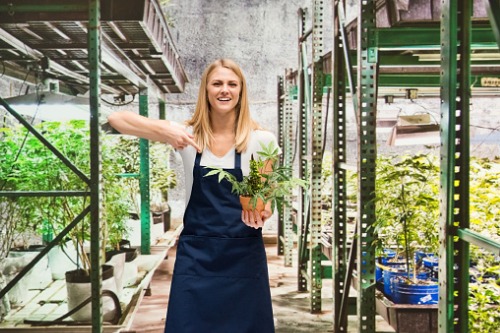 A woman in an apron holding a potted plant in a greenhouse.