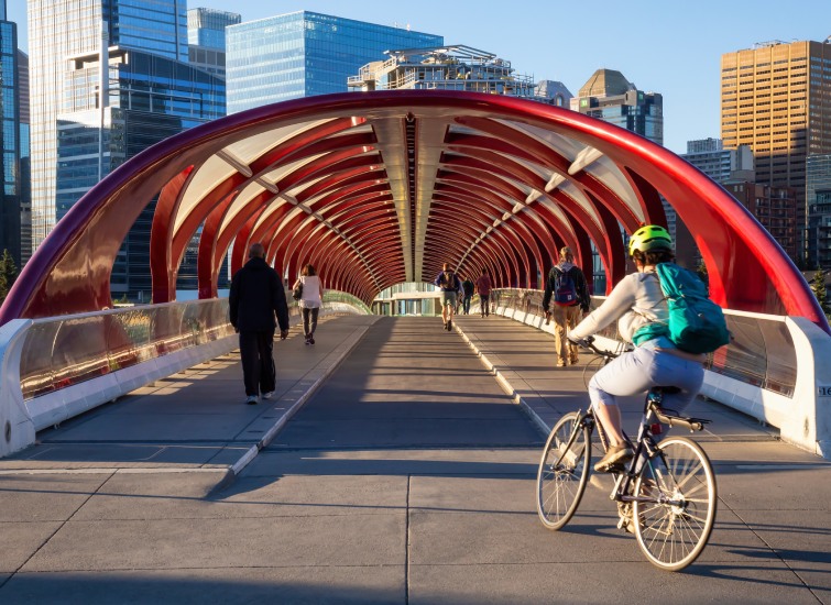 Commuters on bicycles and on foot utilize a modern red pedestrian bridge with a distinctive arch design in an urban setting.