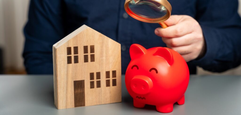 A man with a magnifying glass looking at a piggy bank and a house.