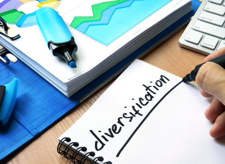 A person writing the word "diversification" on a notebook, with financial charts and a calculator nearby.