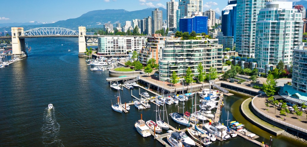 Aerial view of a bustling waterfront district with modern buildings and a marina, bordered by a bridge and mountains in the background.