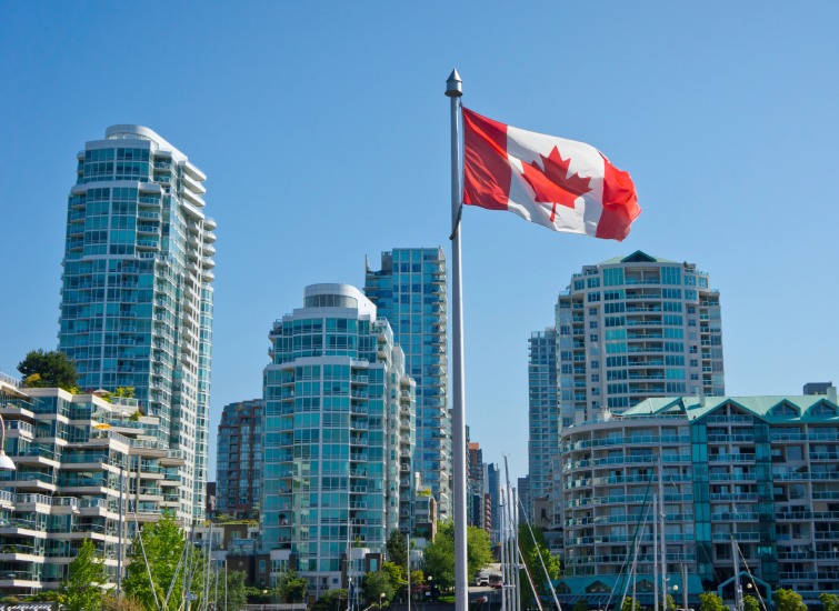 Canadian flag waving in front of modern glass skyscrapers under a clear blue sky.