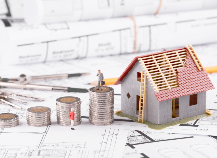 Miniature figure standing on coin stacks next to a small model house on architectural blueprints, symbolizing property investment.