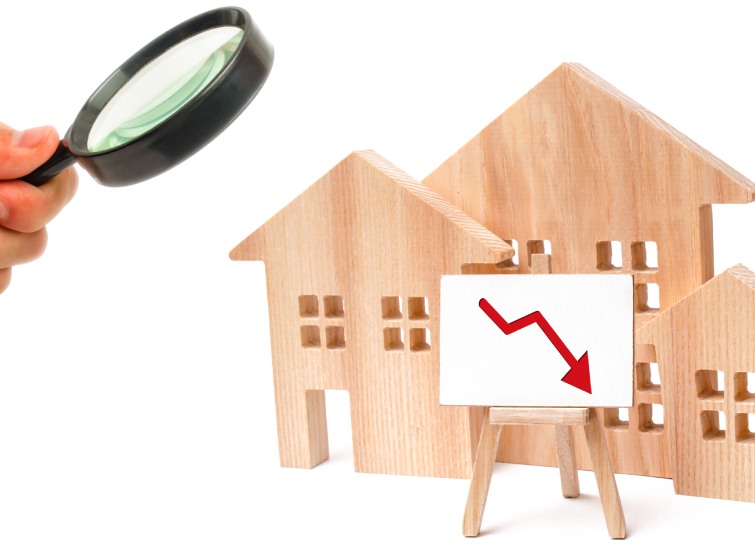 A hand holding a magnifying glass over wooden house models, focusing on one with a downward red arrow indicating a decline in the New Brunswick Real Estate Market.