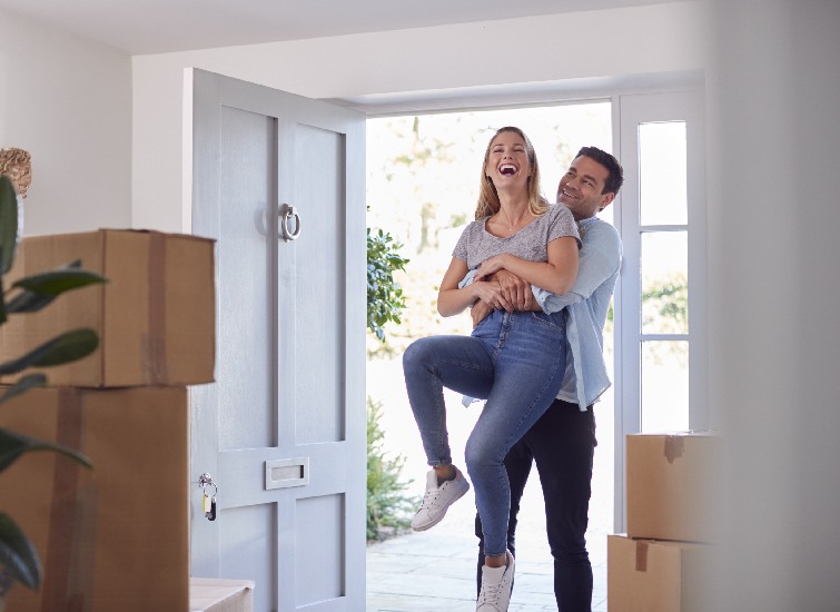 A joyful couple in a new home, with the man carrying the woman across the threshold amid packed moving boxes.