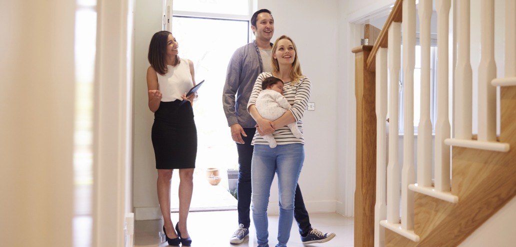A real estate agent showing a couple with a baby around a new home, standing in the hallway and smiling.