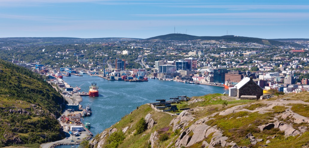 Aerial view of a bustling harbor with ships and cityscape, surrounded by hilly terrain under a clear blue sky.