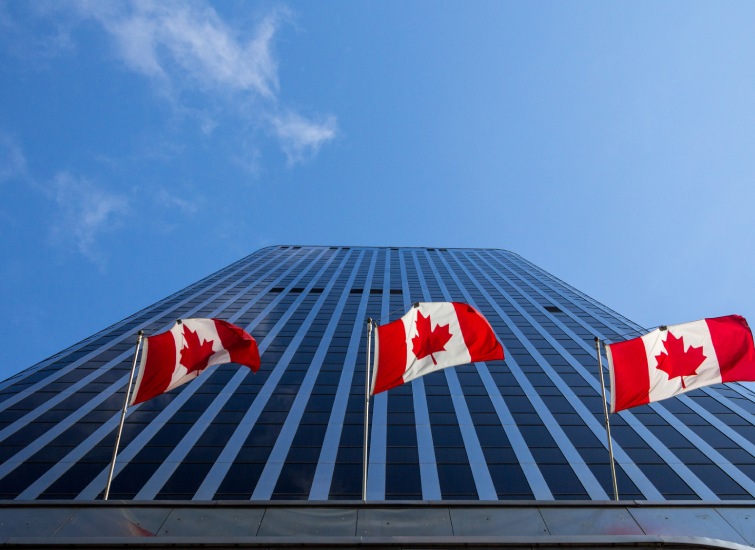 Two Canadian flags waving in front of the Bank of Canada's modern skyscraper under a clear blue sky.