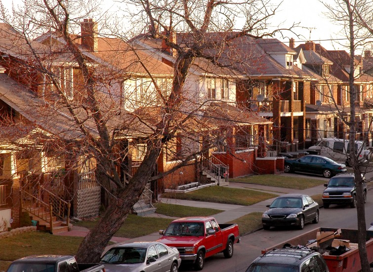 Residential street with parked cars and brick houses at dusk.