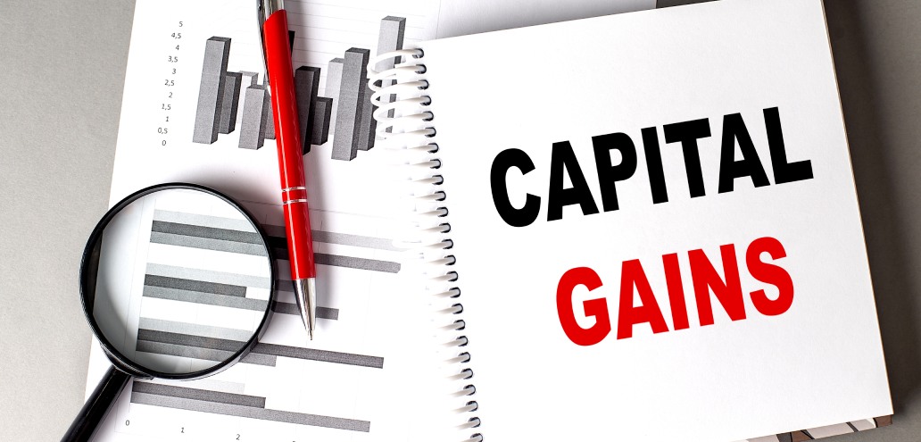 Notebook with "capital gains" text, a magnifying glass, and financial graphs, accompanied by a red pen on a desk.