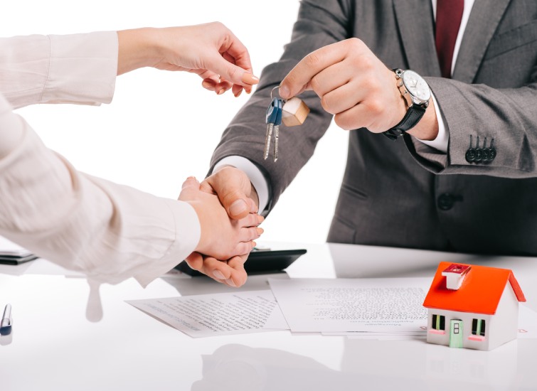 Two people shaking hands over a desk, with one person handing over house keys for a mortgage, and a small red model house on the table.