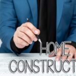 Person in a blue blazer holding a pen sits at a white table with a notebook, coffee cup, and the words "Home Construction" written in front.