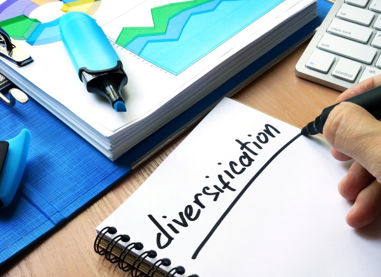 A person writes the word "diversification" on a notepad related to Metro Detroit Real Estate Investments, surrounded by colorful graphs, charts, and a laptop on a wooden desk.