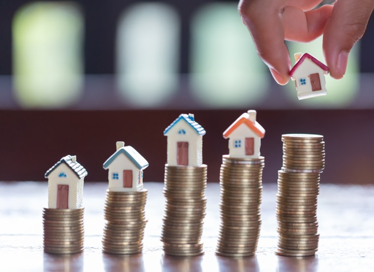 A hand placing a miniature house on top of a stack of coins, symbolizing property investment or real estate value for a revenue property owner.