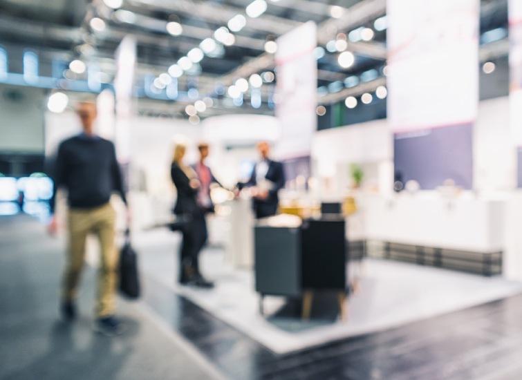 Blurred image of people walking and conversing in a modern, well-lit exhibition hall or trade show with various booths and displays.