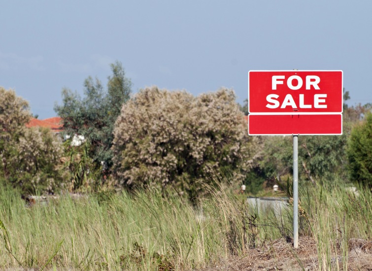 A red "For Sale" sign stands in a grassy, undeveloped lot with trees and a building visible in the background.