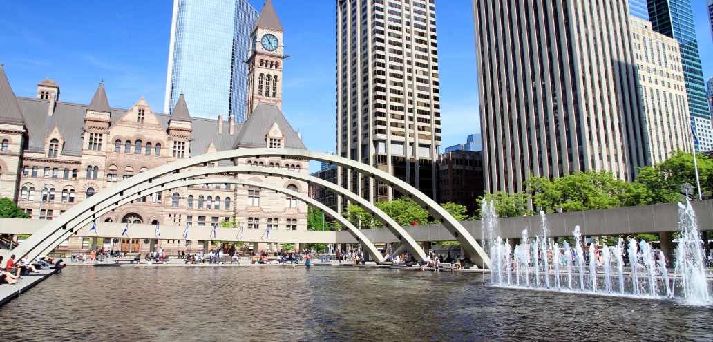 Nathan phillips square in toronto with its iconic arches, fountain, and the old city hall clock tower in the background on a sunny day.