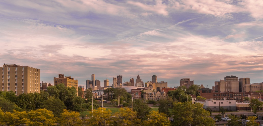 Panoramic view of the Metro Detroit skyline at dusk with scattered clouds and scattered buildings surrounded by trees.