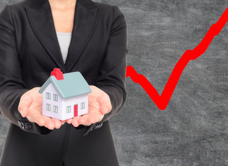 A businesswoman holds a small model house in her hands against a background with an upward trending red graph, symbolizing rising real estate values.