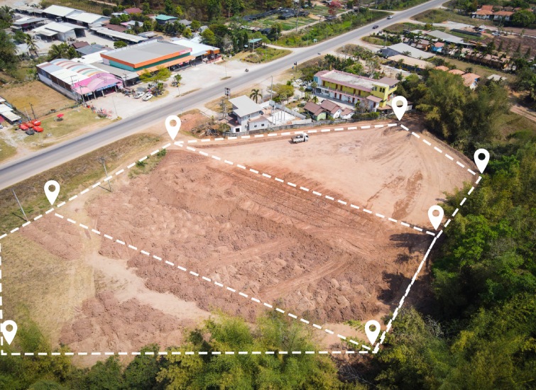 Aerial view of a cleared construction site outlined with a dashed white border and location markers, adjacent to a busy road and various buildings.
